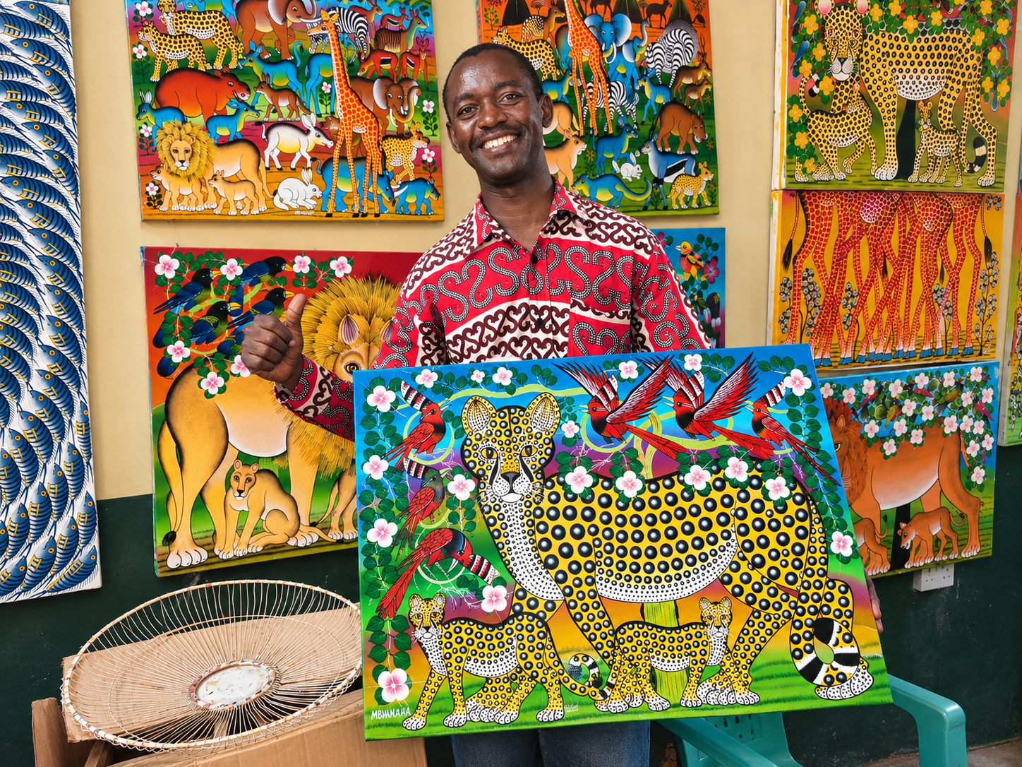 Tanzanian African artist proudly holding a handmade African wildlife painting featuring spotted leopards, cubs, colourful birds and flowers in his art studio surrounded by vibrant African paintings.