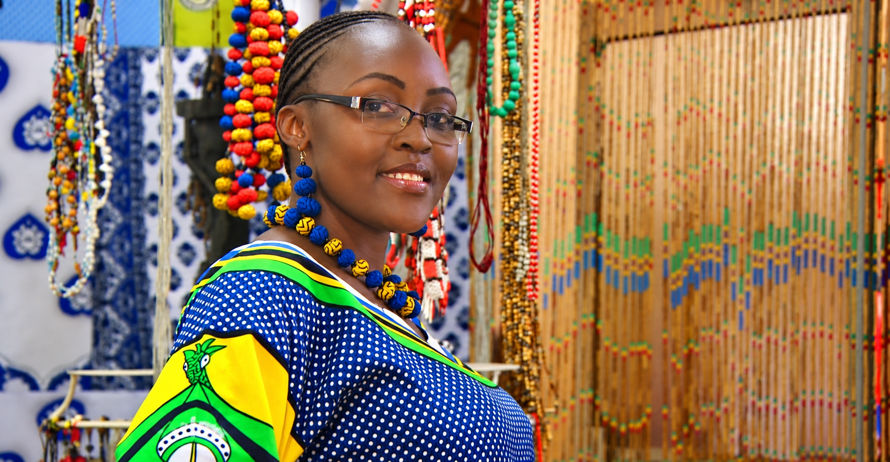 Smiling African artist wearing traditional patterned clothing and handmade beaded jewellery in a colourful African crafts market.